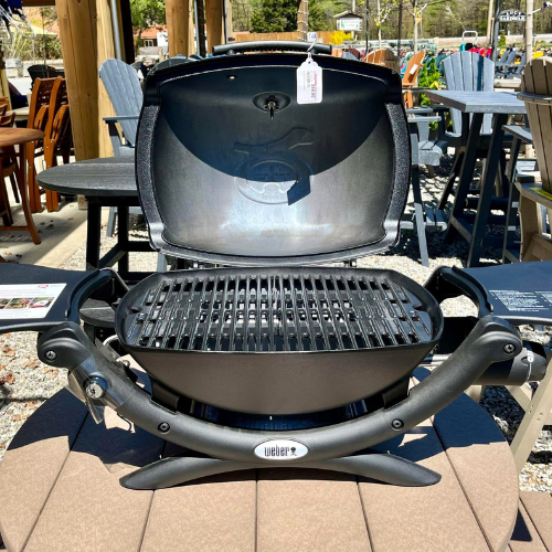 Portable black grill displayed outdoors on a patio, lid open to reveal clean grates. Surrounding seating suggests a relaxed, sunny setting.
