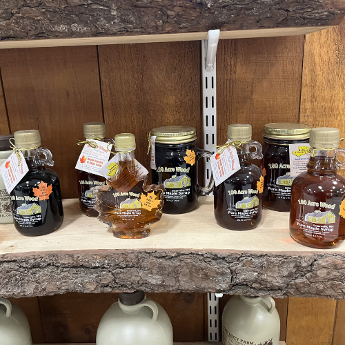 A wooden shelf displays assorted glass bottles of maple syrup with vintage-style labels, offering a rustic, cozy feel. Large ceramic jugs below add charm.