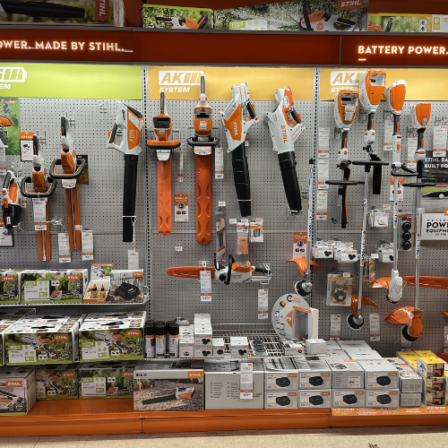 Display of STIHL battery-powered tools on a pegboard wall, including chainsaws, trimmers, and blowers. Boxes on shelves below, showcasing product variety.