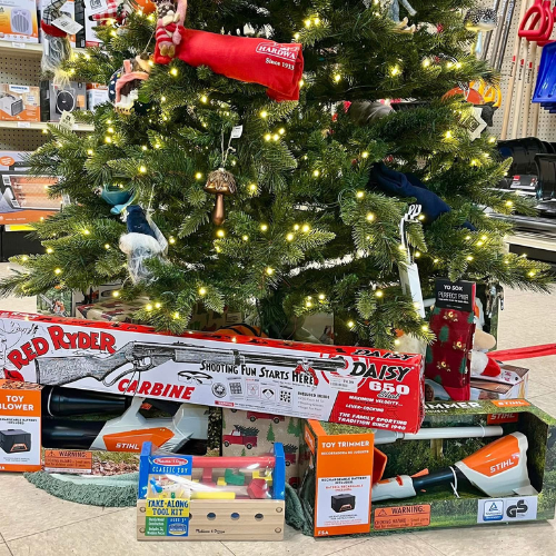 A Christmas tree adorned with lights and ornaments stands in a store. Below, toy guns, tools, and a plush toy are displayed, evoking a festive holiday shopping atmosphere.