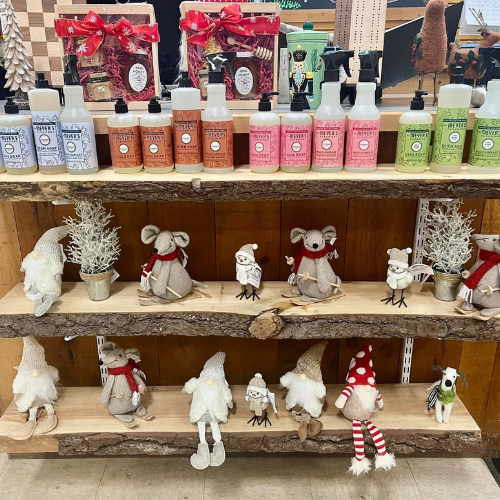 A wooden display shelf with festive gnomes and animal figurines, surrounded by potted plants. Above are colorful bottles of soap, adding charm and coziness.