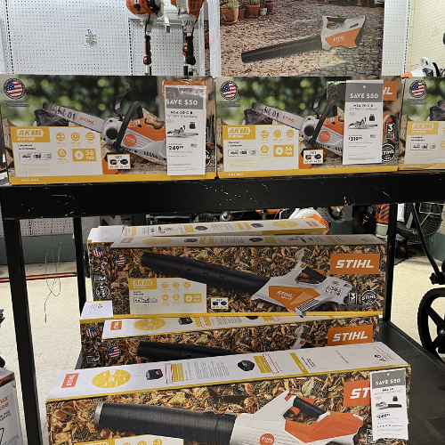 Stacks of STIHL leaf blower boxes on display in a store. The boxes feature images of the blowers and promotional savings offers. Background shows additional tools.