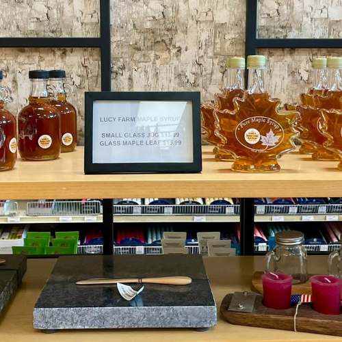 Maple syrup bottles displayed on a wooden shelf, including round and maple leaf-shaped glass jars. A framed sign shows pricing. A rustic scene with warm tones.