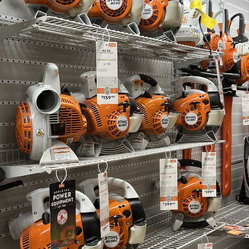 A store display of orange and white Stihl leaf blowers arranged on metal shelves, each with price tags and product information, conveying a practical, organized retail setting.