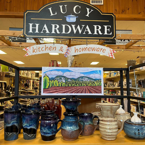 Display in a hardware store featuring kitchenware items like blue and brown pottery mugs and jugs. A colorful landscape painting with swirling clouds is prominently showcased. The atmosphere is cozy and inviting.