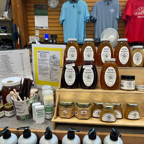 A store display featuring various jars of raw honey on a wooden stand, surrounded by lotions and pain salves. A price list and informational sign are visible, with colorful t-shirts hanging on a wooden wall in the background, creating a quaint, artisanal vibe.
