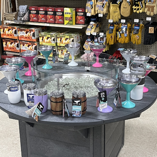 A store display featuring colorful insulated cups, mugs, and tumblers on a circular table. Background shelves are stocked with gloves and fireplace supplies.