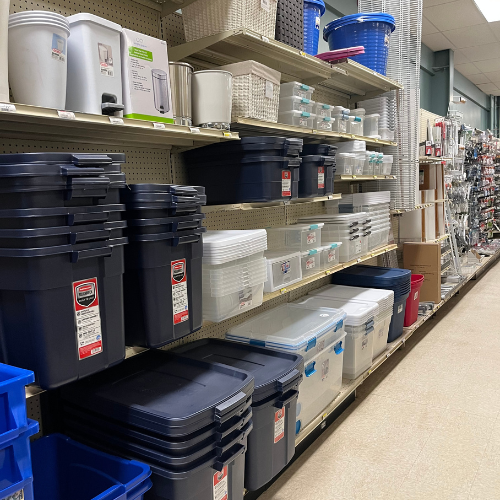Store aisle with organized shelves displaying various plastic storage bins in blue, white, and clear. Shelving units also hold baskets and containers.