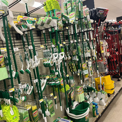 A store aisle displays cleaning supplies, including green and white brushes, mops, gloves, and buckets. The area is neatly organized, conveying a sense of cleanliness.