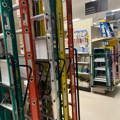A hardware store aisle featuring various ladders in green, red, and yellow. Nearby shelves display paintbrushes and cleaning supplies. Brightly lit, organized.