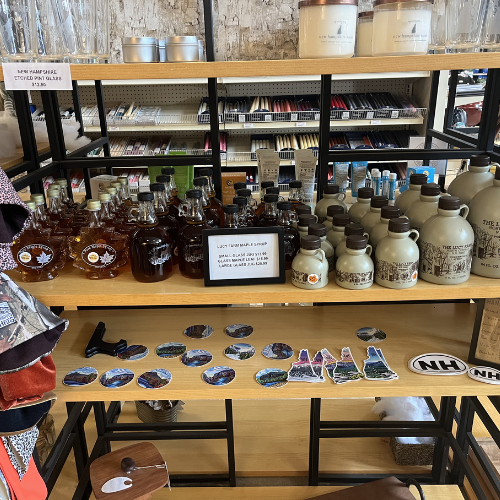 Shelves display maple syrup bottles in glass and ceramic jugs, stickers with landscapes, and candles. Cozy, rustic shop ambiance.