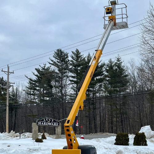 A tall yellow lift extends upwards beside a snow-covered hardware store sign with trees and cloudy skies in the background, conveying a wintery, industrious scene.