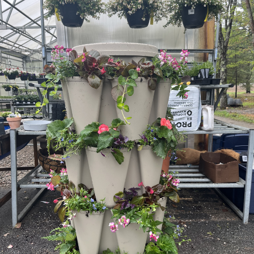 Vertical planter tower in a greenhouse, adorned with vibrant flowers and leafy plants. Background includes shelves of additional greenery, creating a lush, lively atmosphere.