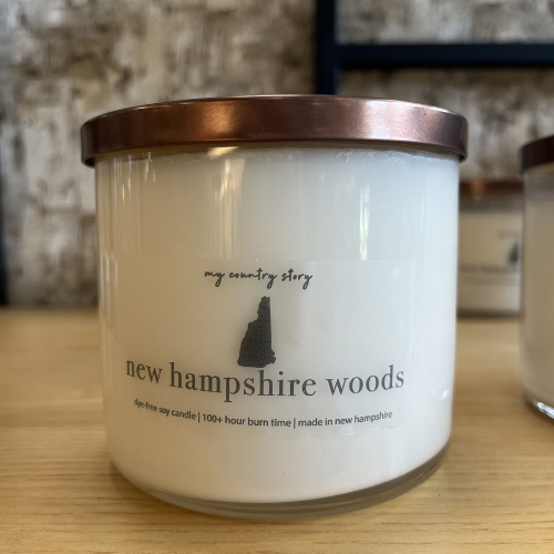 A white candle in a glass jar with a copper lid sits on a wooden surface. The label reads 