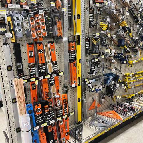 A hardware store aisle displays various measuring tools. Bright orange, yellow, and silver levels and squares are neatly arranged on pegboards, evoking a sense of order and utility.