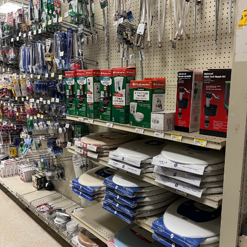 Hardware store aisle with shelves of plumbing supplies, including packaging for toilet seats, repair kits, and various pipe fittings. Functional atmosphere.