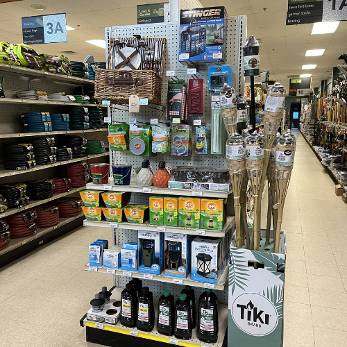 An in-store display of outdoor products, including tiki torches, insect repellents, and citronella candles. Store aisles stocked with hoses and gardening tools.