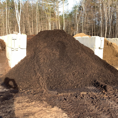 A large, dark brown pile of mulch is placed between concrete barriers, surrounded by trees under a clear sky, suggesting a natural, outdoor setting.