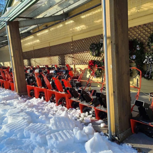 Red snow blowers neatly lined up under a wooden shelter, surrounded by snow. The setting is bright and festive with decorated wreaths on the wall.