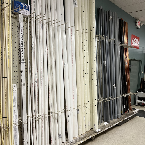 Rows of metal racks hold various long, cylindrical pipes in white, gray, black, and copper hues against a store's light green wall, evoking an industrial vibe.