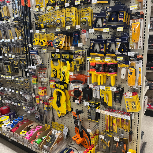 A hardware store display shelf features various tools and accessories. Bright yellow and orange packaging stands out, showcasing brands like Stanley. The shelves are filled with measuring tapes, utility knives, and duct tape rolls. The organized layout conveys a sense of readiness and practicality.