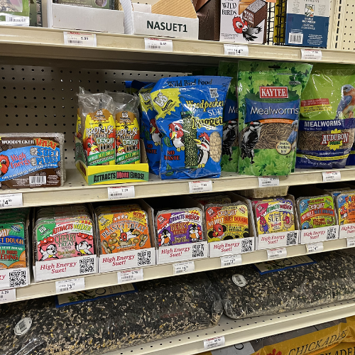 Store shelf displaying various bird feed products. Colorful packaging includes suet cakes, seed, and mealworms. Labels emphasize energy and attracting birds.