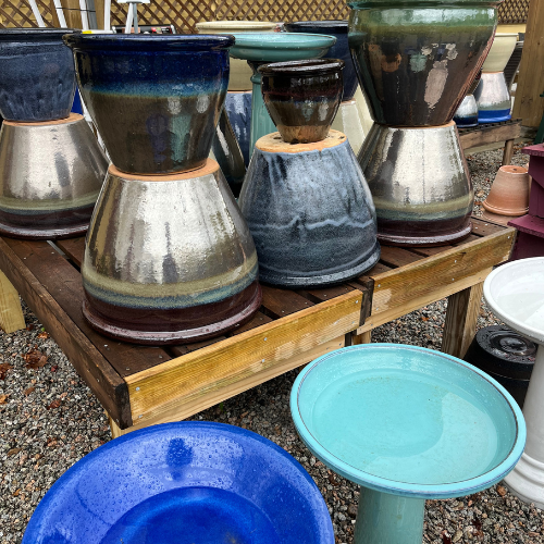 Ceramic planters and bird baths in earthy tones and vibrant blues are displayed on wooden shelves. The setting is outdoors with a gravel surface.