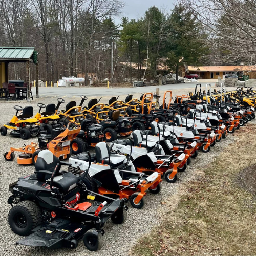 A lineup of various lawnmowers on gravel, featuring bright orange and black designs. Trees and low buildings are visible in the background.
