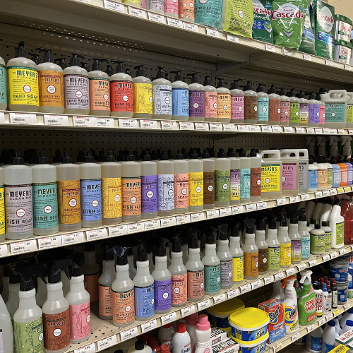 Shelves of colorful cleaning products, including Mrs. Meyer's soap in pastel bottles, neatly arranged. Bright, organized, and inviting display.