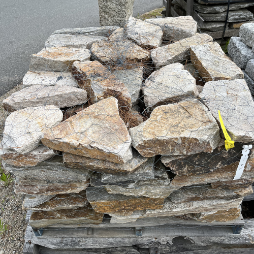 A pallet stacked with variously shaped, rough-cut stones secured by netting. The stones are earthy-toned; a yellow tag is attached to one.
