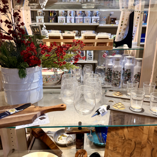 A display shelf in a store with festive decor. Features clear glasses, vibrant red berries in a white pot, cutting boards, and floral bottles, creating a cozy, inviting feel.