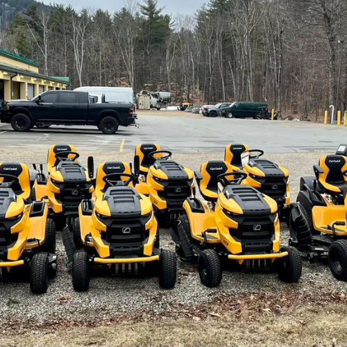 A row of vibrant yellow lawn tractors is arranged in a parking lot with a forest backdrop. Nearby are parked vehicles, including a black truck.