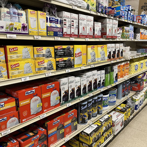 Store shelves stocked with colorful packaging of various brands of trash bags and paper towels. The aisle appears well-organized and brightly lit.