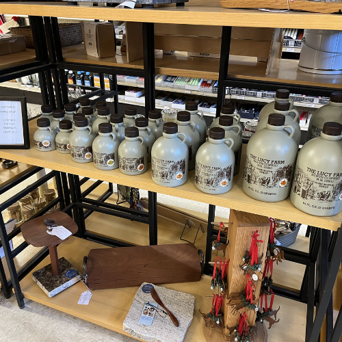 Shelves display gray jugs of The Lucy Farm syrup with black caps. Below are wooden cutting boards, a knife, and red tassel ornaments, creating a rustic, cozy vibe.