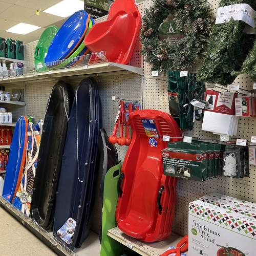 Store aisle with colorful sleds, wreaths, and Christmas decorations on shelves. Festive and playful atmosphere with holiday-themed products.