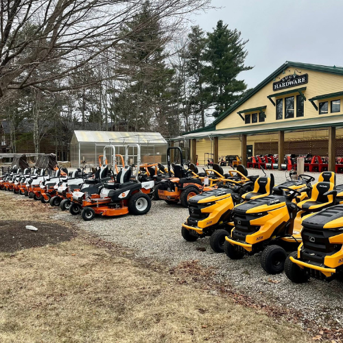 A row of yellow and orange riding lawn mowers displayed outside a hardware store beneath bare trees, conveying a sense of readiness for spring.