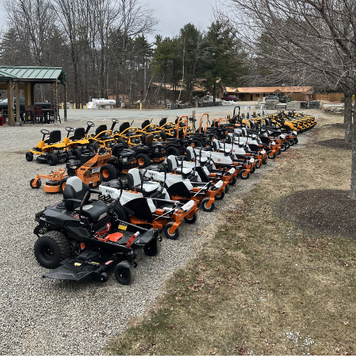 A row of various lawn mowers displayed on gravel outside, with bare trees and buildings in the background. The scene feels organized and ready for sale.