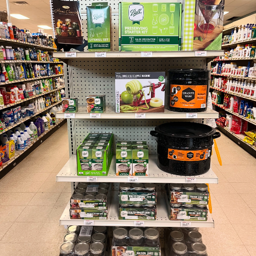 Store aisle display featuring canning supplies, apple peelers, and a large pot. Shelves are stocked neatly with vibrant packaging, conveying a sense of organization.