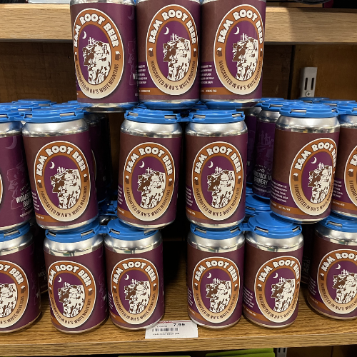 Cans of Ikar Root Beer are stacked on a wooden shelf. Each can is purple with a circular logo featuring a ship in a storm, conveying a nautical theme.