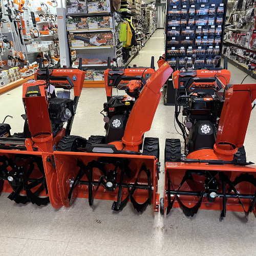 Three orange snow blowers are displayed in a hardware store aisle. Shelves behind them are stocked with tools and equipment, conveying a sense of preparedness.