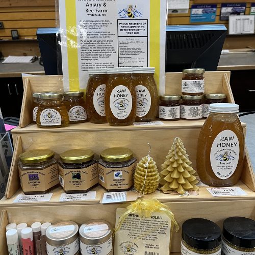 Wooden display shelves hold jars of raw honey, honeycomb, and beeswax candles in a farm shop. Labels highlight local production. Cozy and rustic ambiance.