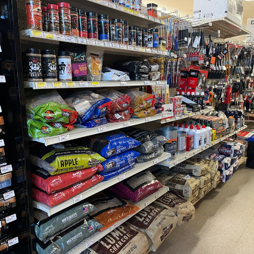 A store aisle filled with barbecue supplies, including shelves stocked with charcoal bags, wood pellets, grilling tools, and barbecue sauces. The atmosphere is organized and inviting for grilling enthusiasts.