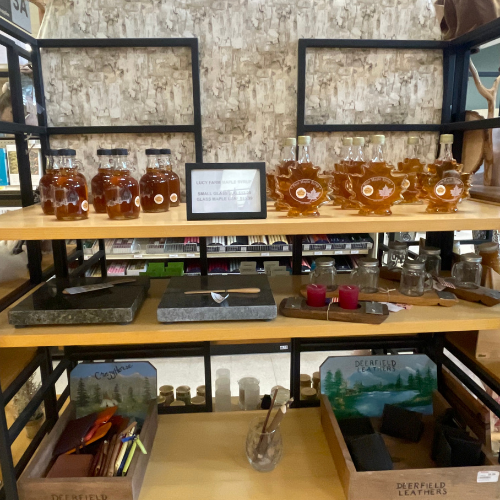 Shelves displaying maple syrup bottles shaped like leaves, stone trays, leather goods, and candles. The scene conveys a rustic, artisanal vibe.