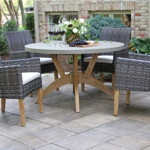 Outdoor patio with a round table set, stone tiles, and comfy wicker chairs. Table adorned with wine glasses, flowers, and a cheese platter; serene, inviting vibe.