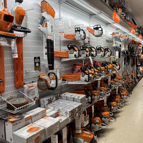 A store aisle displaying a variety of orange and white power tools, including chainsaws and trimmers, neatly arranged on metal shelves.